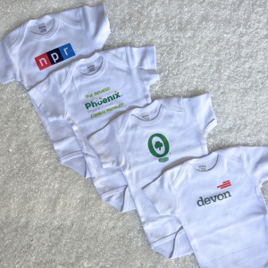 Four white baby onesies spread on a light carpet, each featuring a different logo: NPR, 'The newest Phoenix Physical Therapy family member', a green circular logo with a shamrock, and 'devon' with a red bar icon.